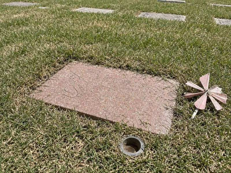 Illustrative example, before: a small pink granite infant marker with an angel etching, grass over its edges and a faded pinwheel